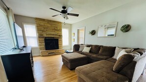 A living room with a stone fireplace, a large sectional sofa, a ceiling fan, and a flat-screen TV. - Charming 3-bedroom house in historic grandview neighborhoods  (Dubuque)