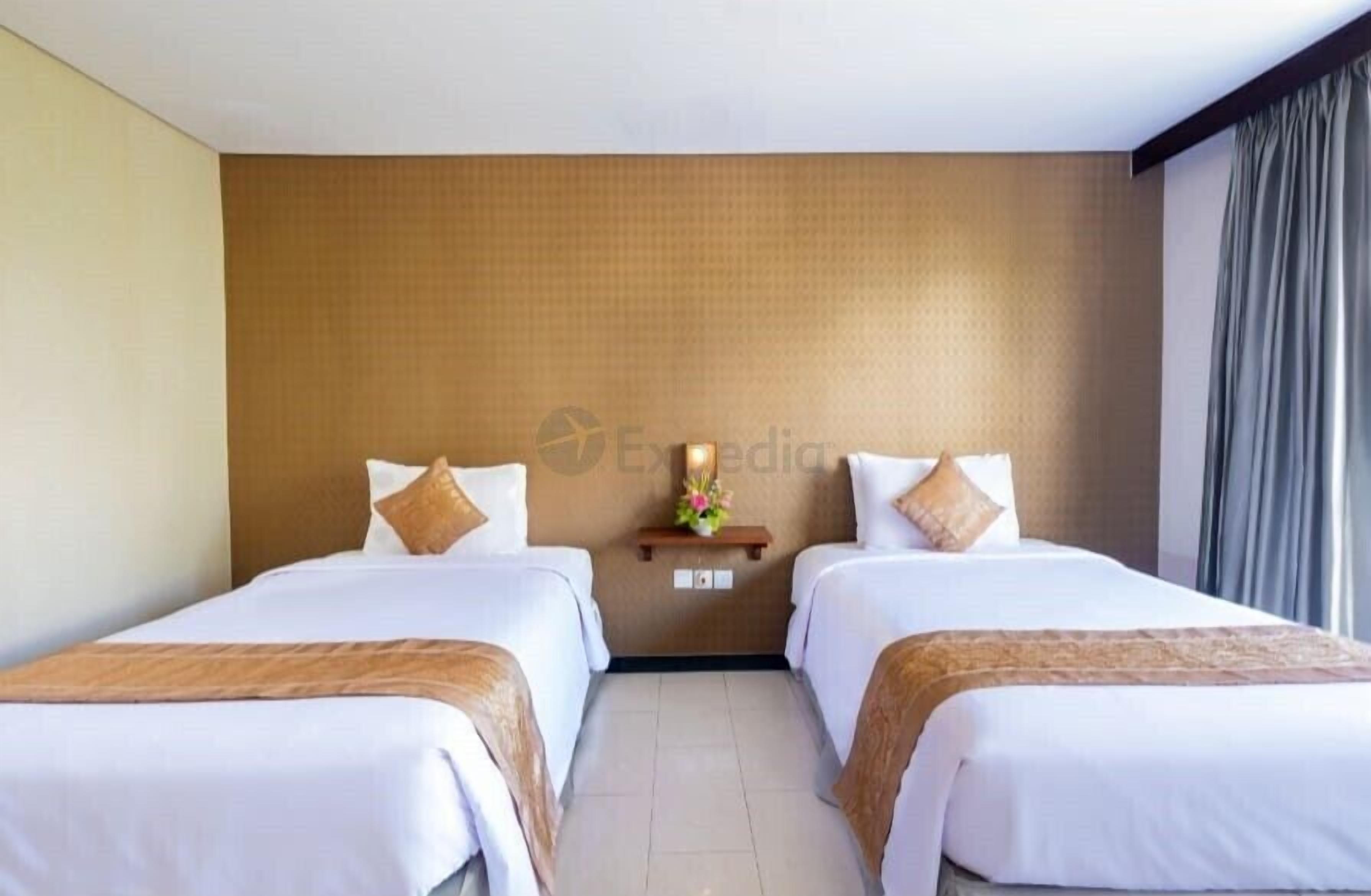 A hotel room with two beds, a wooden headboard, a small shelf with a vase, and a window with curtains.
