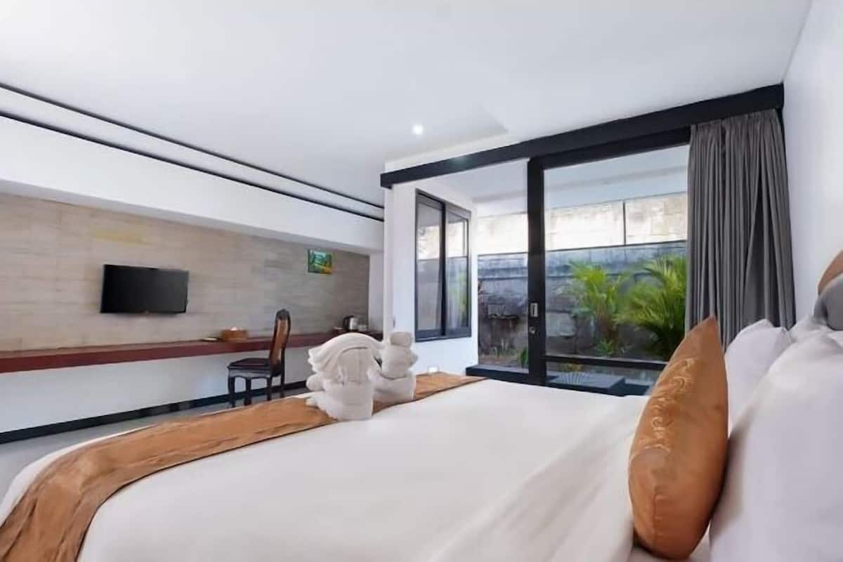 A modern hotel room with a large bed, a flat-screen TV, a view of greenery, and a sliding door to the outside.