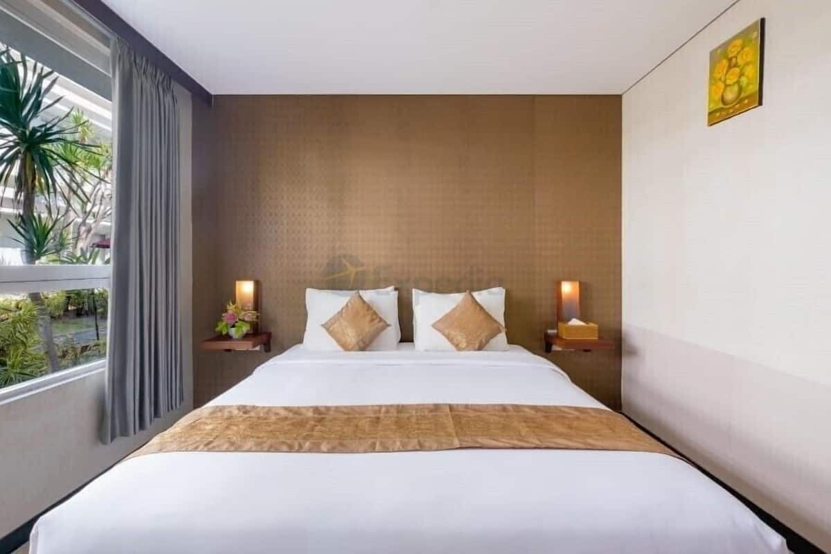 A hotel room with a large bed, two bedside tables with lamps, a window with curtains, and a view of greenery outside.