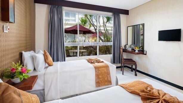A hotel room with two beds, a desk, a chair, a TV, and a window with a view of palm trees and a building.