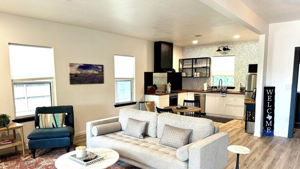A modern living room with a sofa, coffee table, and patterned rug. A kitchen is visible in the background with a welcome sign. - ꕥ The Texas Bluebonnet ꕥ 1.1 mi to Baylor, & 1.8 mi to Magnolia Silos! (Waco)