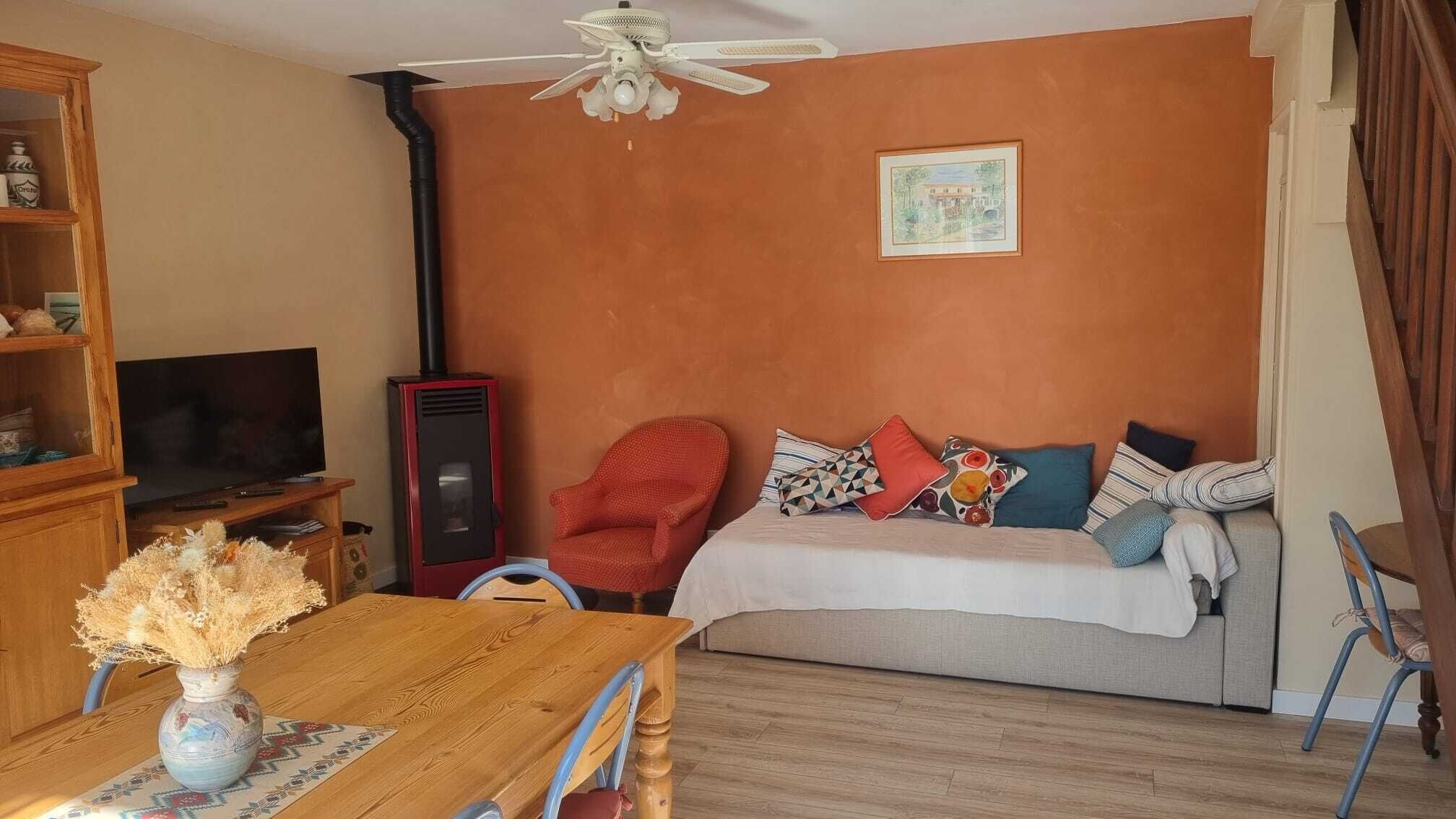 A cozy living room with a wooden table, a red armchair, a sofa with colorful pillows, a ceiling fan, and a TV.