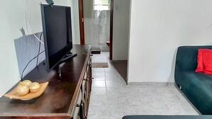 A living room with a wooden TV stand, a flat-screen TV, a wooden table with a plate of fruit, a teal sofa with a red pillow, and a tiled floor.