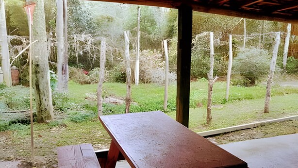 A covered outdoor seating area with a wooden bench and table, surrounded by a garden with trees and a fence.
