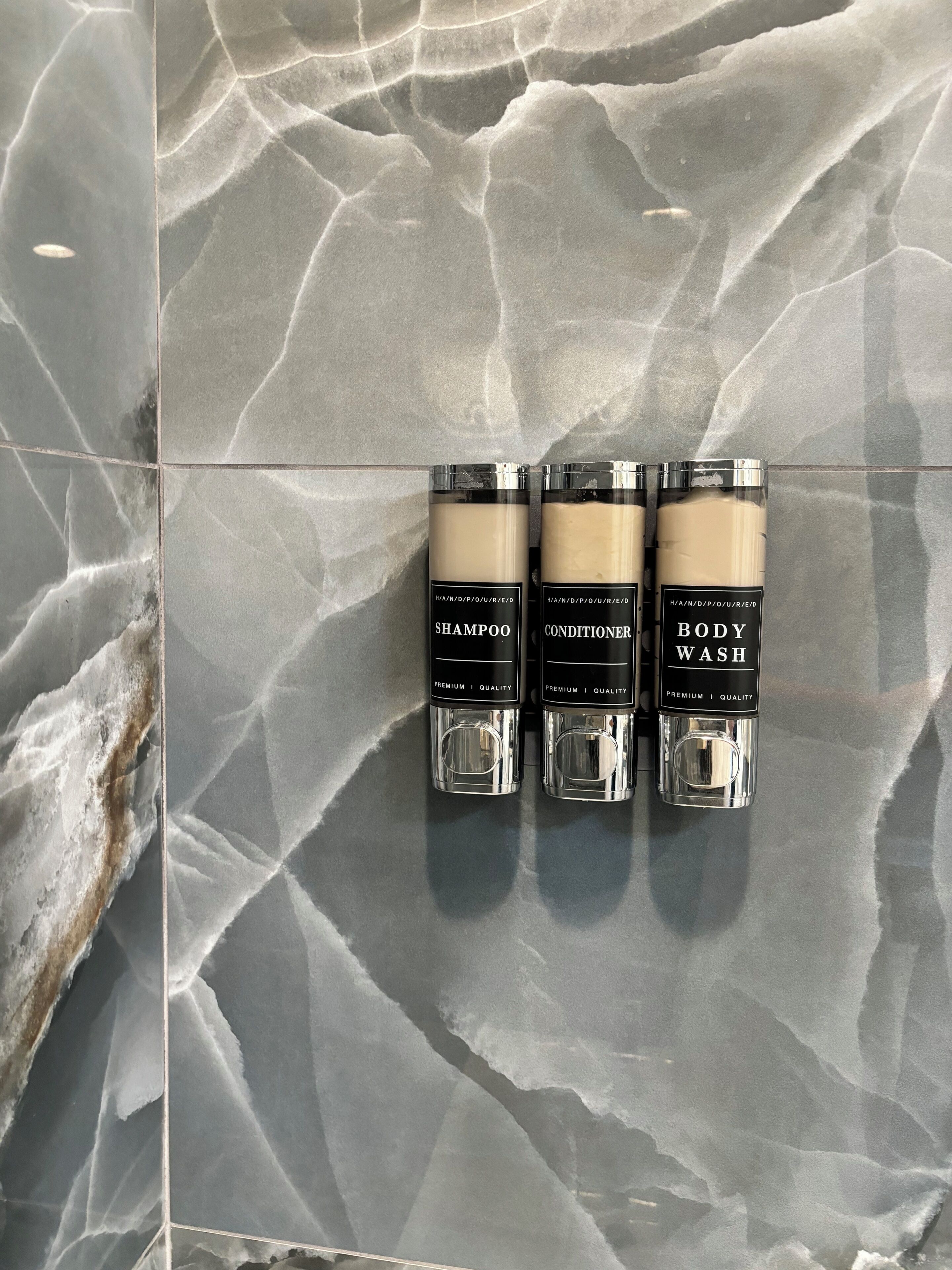 Three toiletry bottles labeled 'SHAMPOO', 'CONDITIONER', and 'BODY WASH' on a tiled wall.