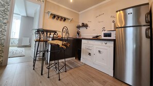 Une cuisine moderne équipée d’un réfrigérateur en acier inoxydable, d’un four à micro-ondes et d’une machine à café posés sur le plan de travail. On y trouve deux chaises en métal noir avec des coussins, une petite table et un tapis à motifs sur le sol.