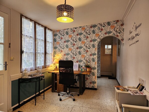 Une pièce comprenant un bureau, une chaise et une lampe. On y trouve des fenêtres avec des rideaux, un mur recouvert d’un papier peint à motifs floraux et une porte.