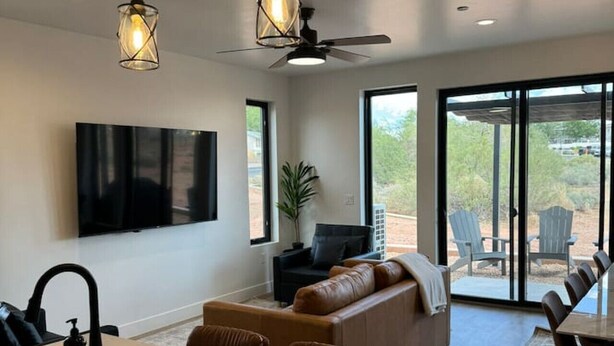Et moderne køkken med en central ø, et spiseområde med bord og stole, en opholdsstue med sofa, og et stort vindue med udsigt til naturen.