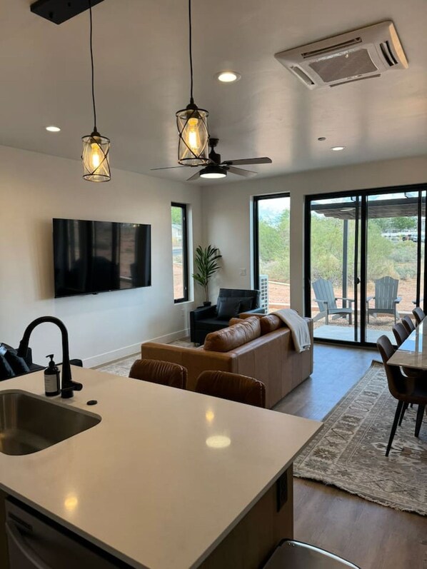 A modern kitchen with a central island, a dining area with a table and chairs, a seating area with a sofa, and a large window offering a view of the outdoors.