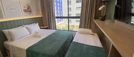A hotel room with two beds, a desk, a window with curtains, and artwork on the walls.