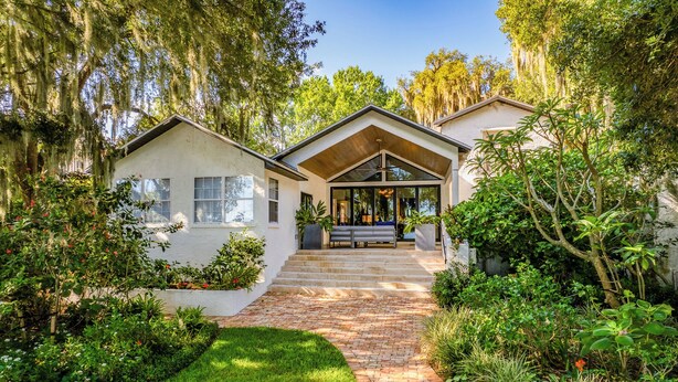 A house with a covered porch, a brick pathway leading to the entrance, and lush greenery surrounding the property. - Elegant Lake Retreat (Mount Dora)