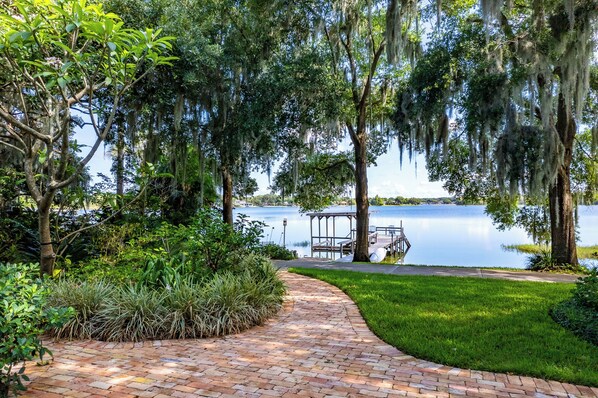 A brick pathway leading to a dock by a calm lake, surrounded by lush greenery and trees with hanging moss.