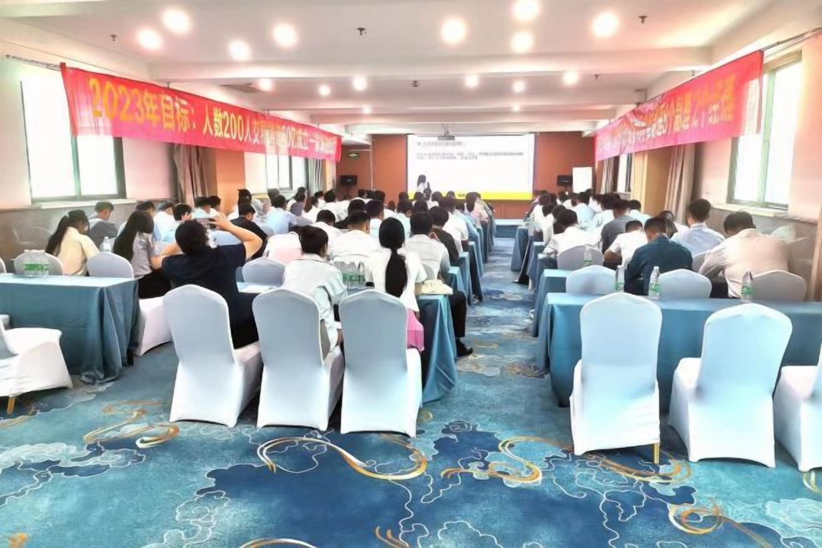 A conference room with rows of tables and chairs, a carpeted floor, and a presentation screen at the front.