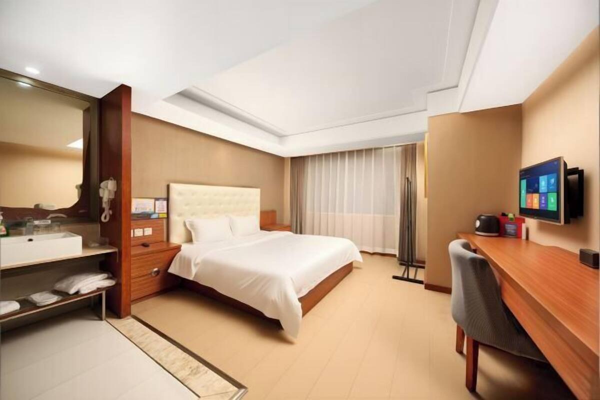 Habitación de hotel con cama, escritorio, silla, televisor, espejo, estante con toallas y ventana con cortinas.