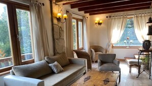 Un salon chaleureux avec un canapé, des fauteuils, une table basse en bois et une vue sur l’extérieur grâce à de grandes fenêtres.