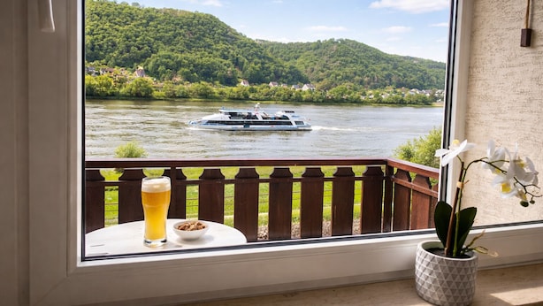 Vista desde una ventana sobre un río con un barco, un balcón con una mesa y una cerveza en un vaso, y una orquídea en maceta.