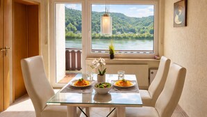 Un comedor con mesa de vidrio, sillas blancas y vista a un lago y bosque.