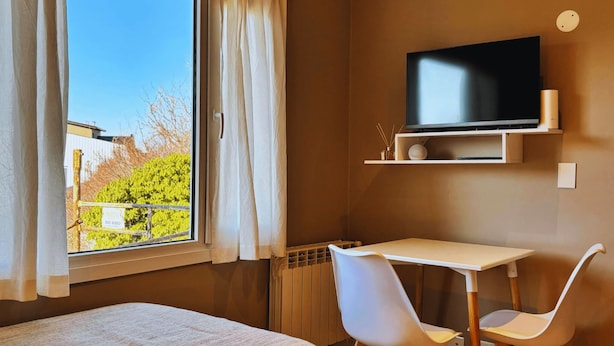 Ein Schlafzimmer mit einem Bett, einem Schreibtisch, zwei Stühlen, einem Fernseher und einem Fenster mit Vorhängen.
