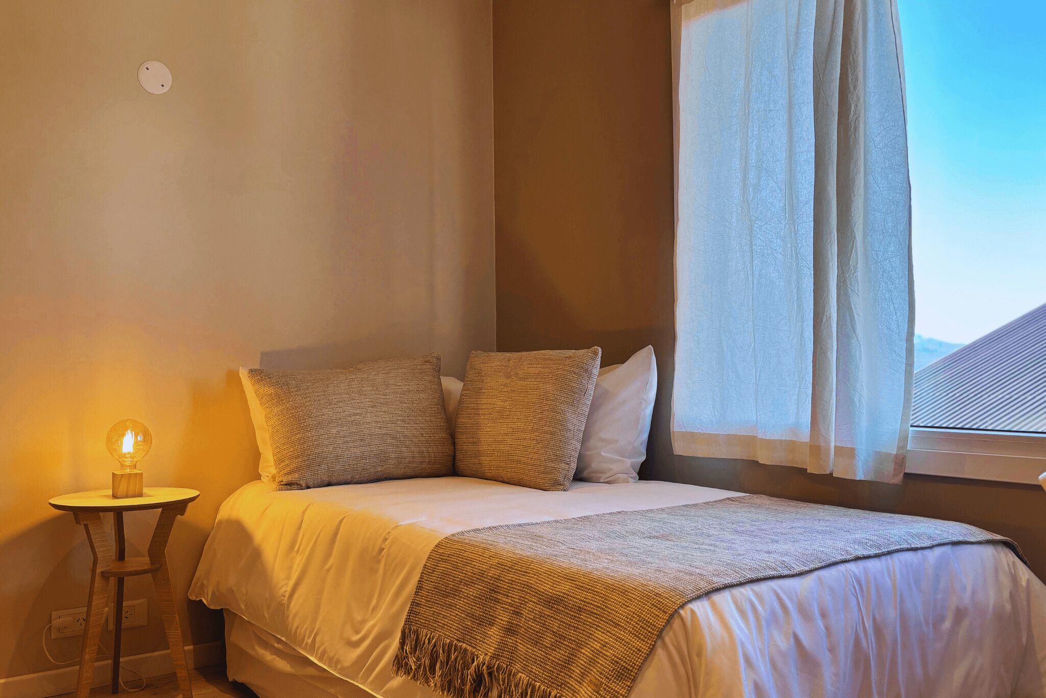 A bedroom with a bed, pillows, a bedside table with a lamp, and a window with curtains.