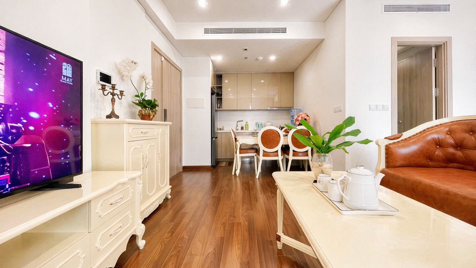 A modern living room with a television, a sofa, a dining area with chairs and a table, and a kitchen in the background.