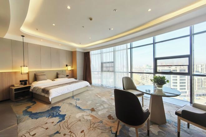 Una habitación de hotel moderna con una cama grande, un escritorio y vistas de la ciudad a través de amplias ventanas.