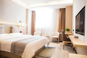 A modern hotel room with a large bed, a desk, two chairs, a TV, and a window with curtains.