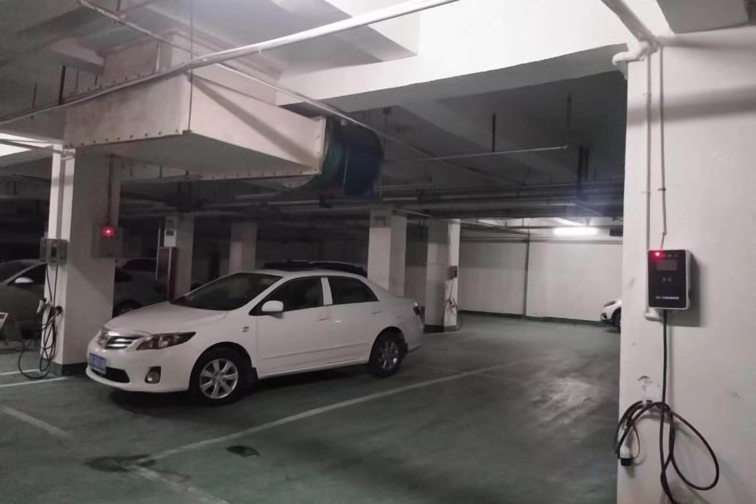 Un auto blanco estacionado en un estacionamiento subterráneo con elementos estructurales visibles y un panel de control en la pared.