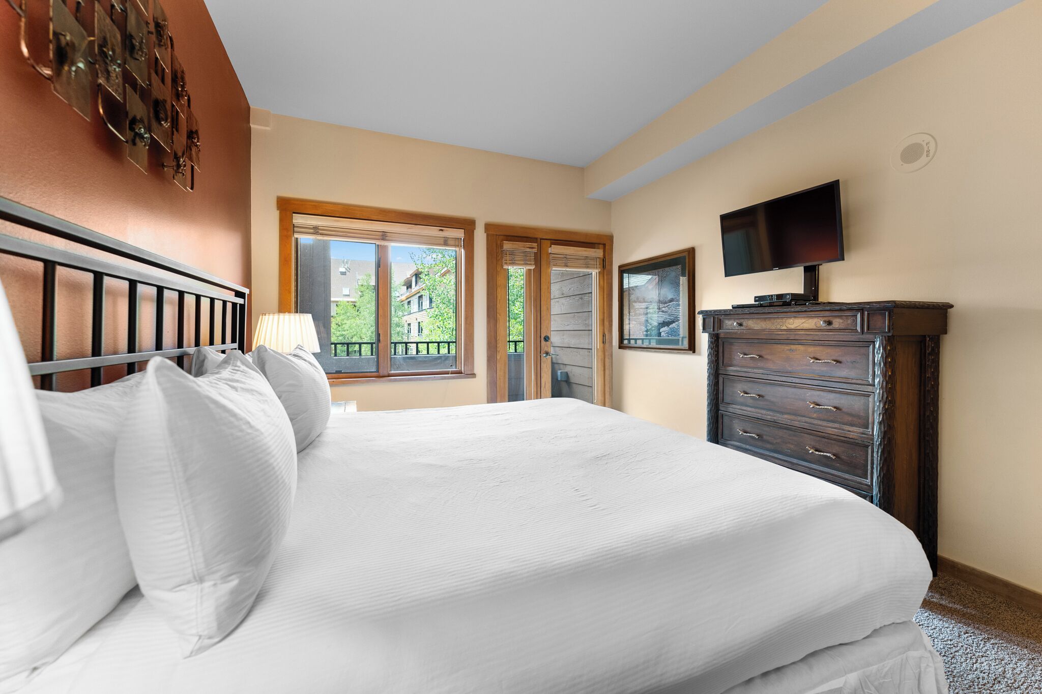 A bedroom with a bed, dresser, TV, and windows with a view of buildings and trees.