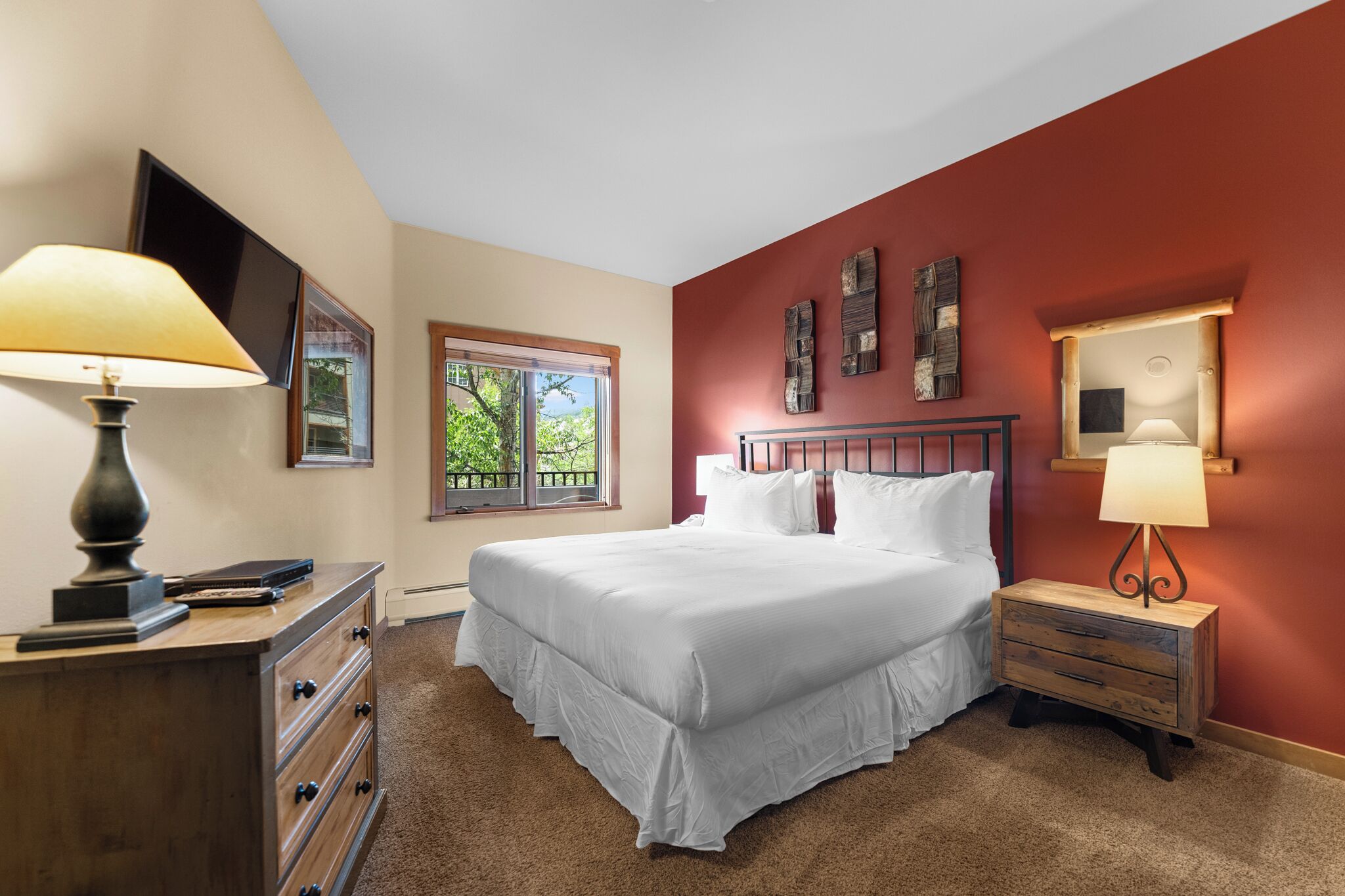 A bedroom with a large bed, a dresser, a nightstand, a TV, and a window with a view of trees.