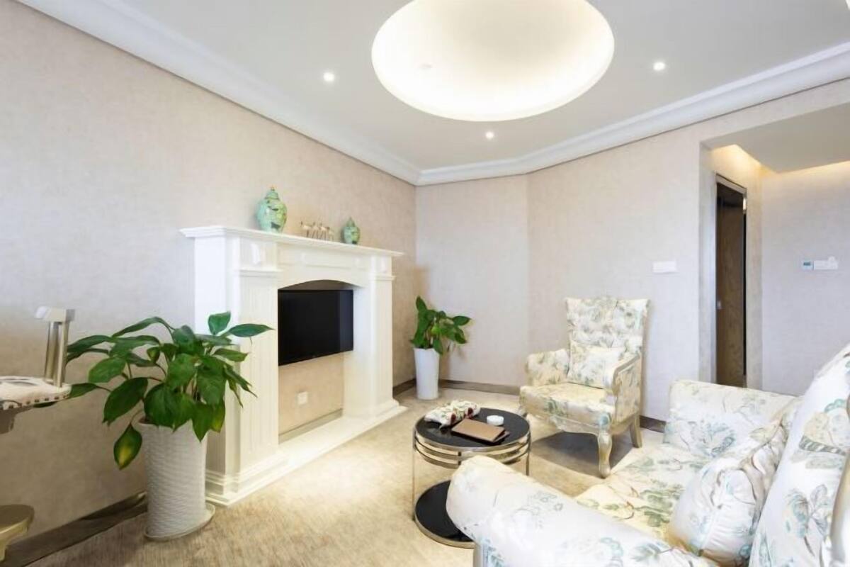 Una sala de estar con chimenea, sillones de estampado floral, una mesa de centro redonda y una planta grande en maceta.