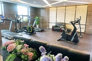 Een sportschool met loopbanden, een elliptische trainer en een hometrainer. Er staat een zwarte leren bank en een potplant met bloemen.