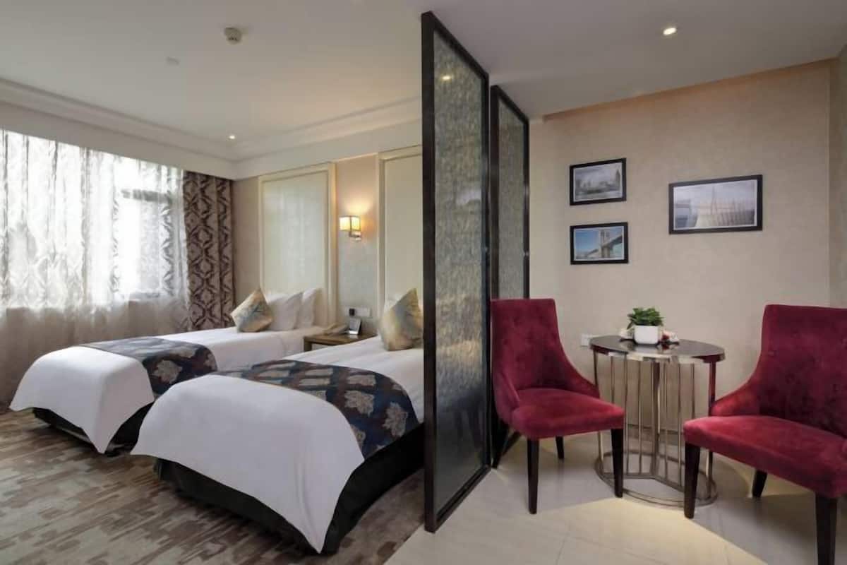 Una habitación de hotel con dos camas, una zona de estar con una mesa y sillas, y un ventanal con cortinas.