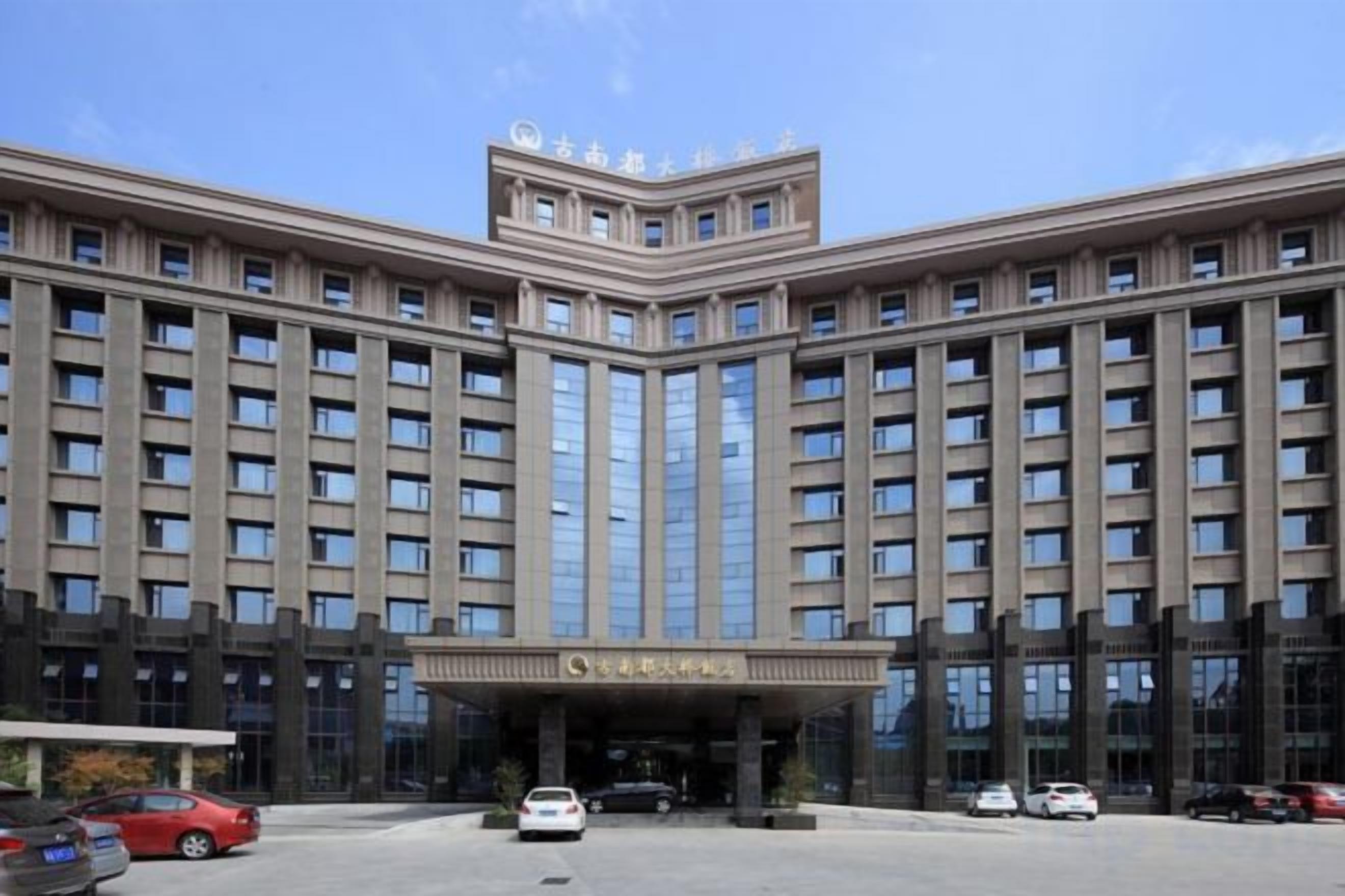 Un edificio grande de varios pisos con fachada simétrica, entrada central y estacionamiento al frente.