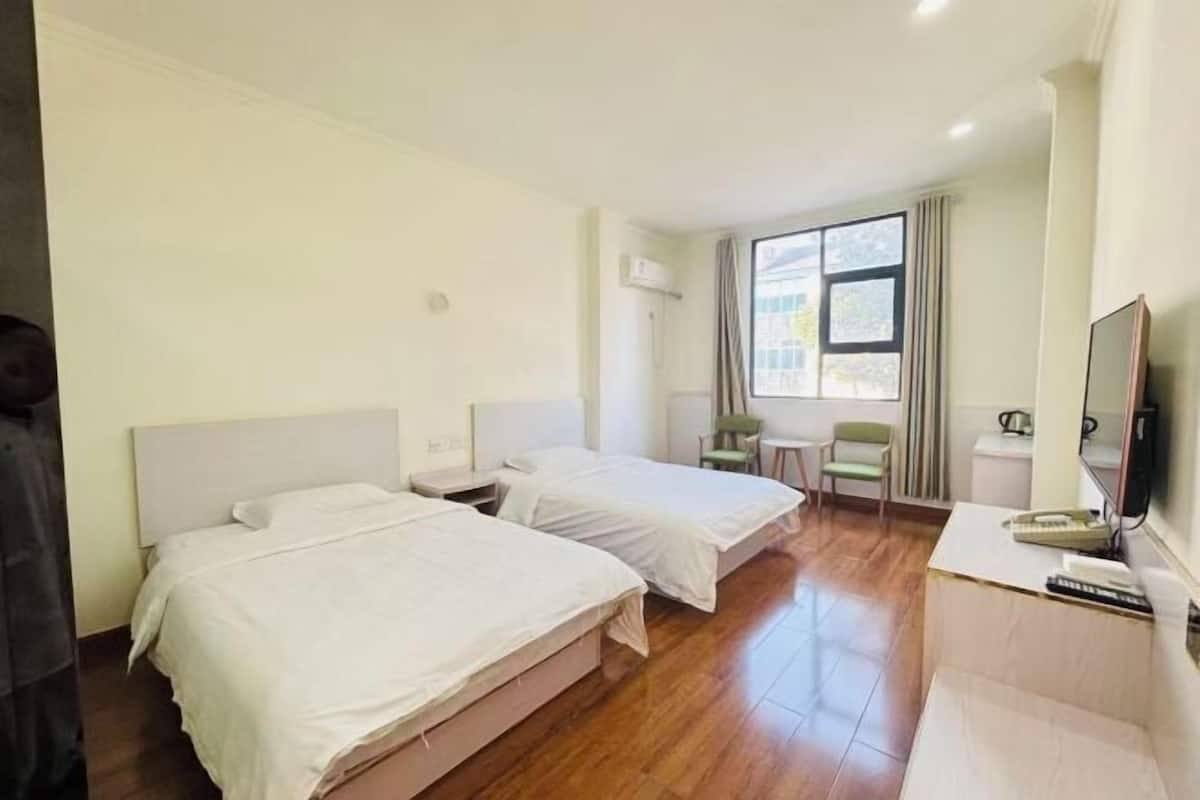 A hotel room with two beds, a small table, a chair, a TV, and a window with curtains.