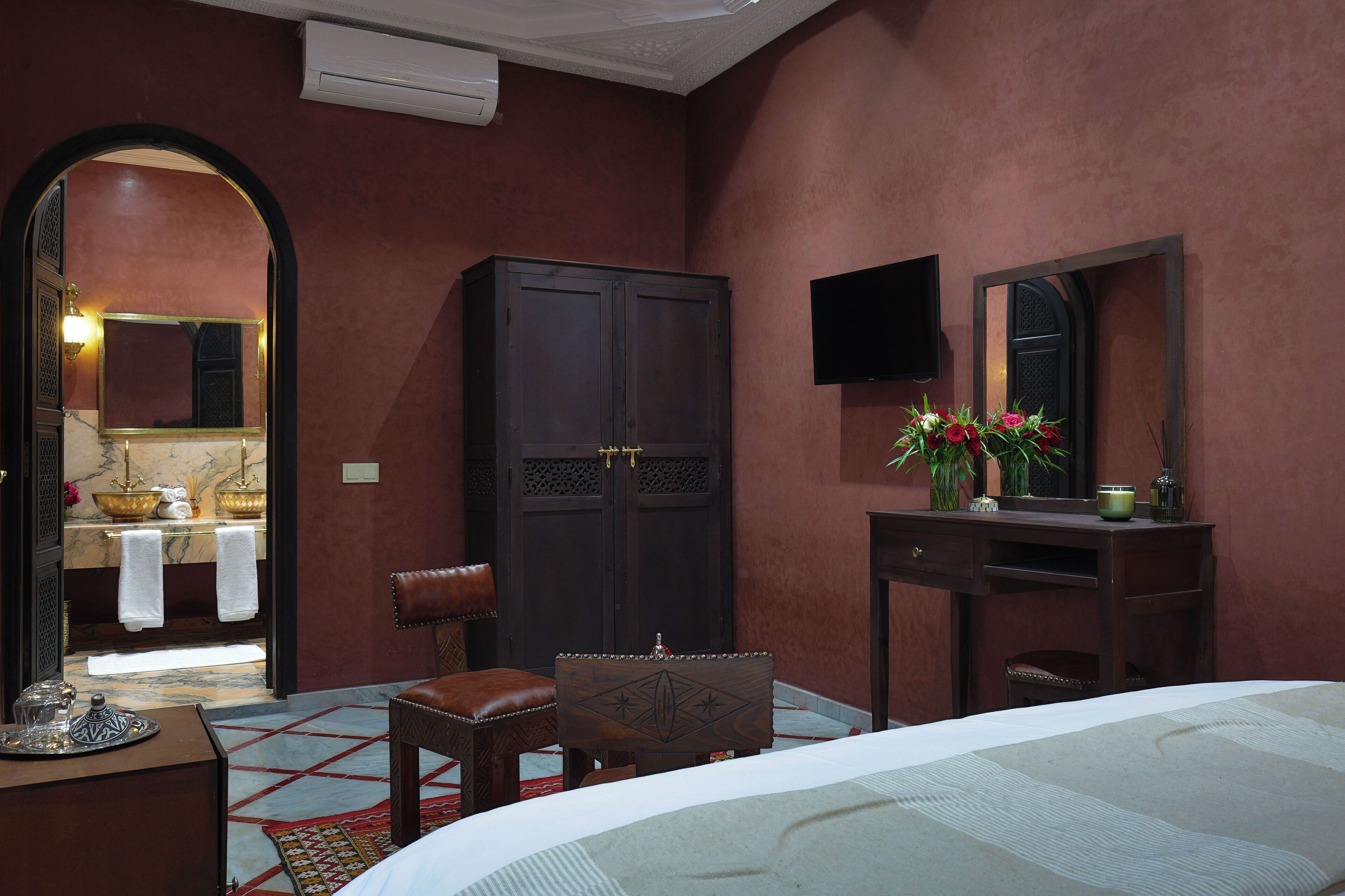 A bedroom with a bed, a desk, a chair, a wardrobe, a TV, and a bathroom visible through an archway.