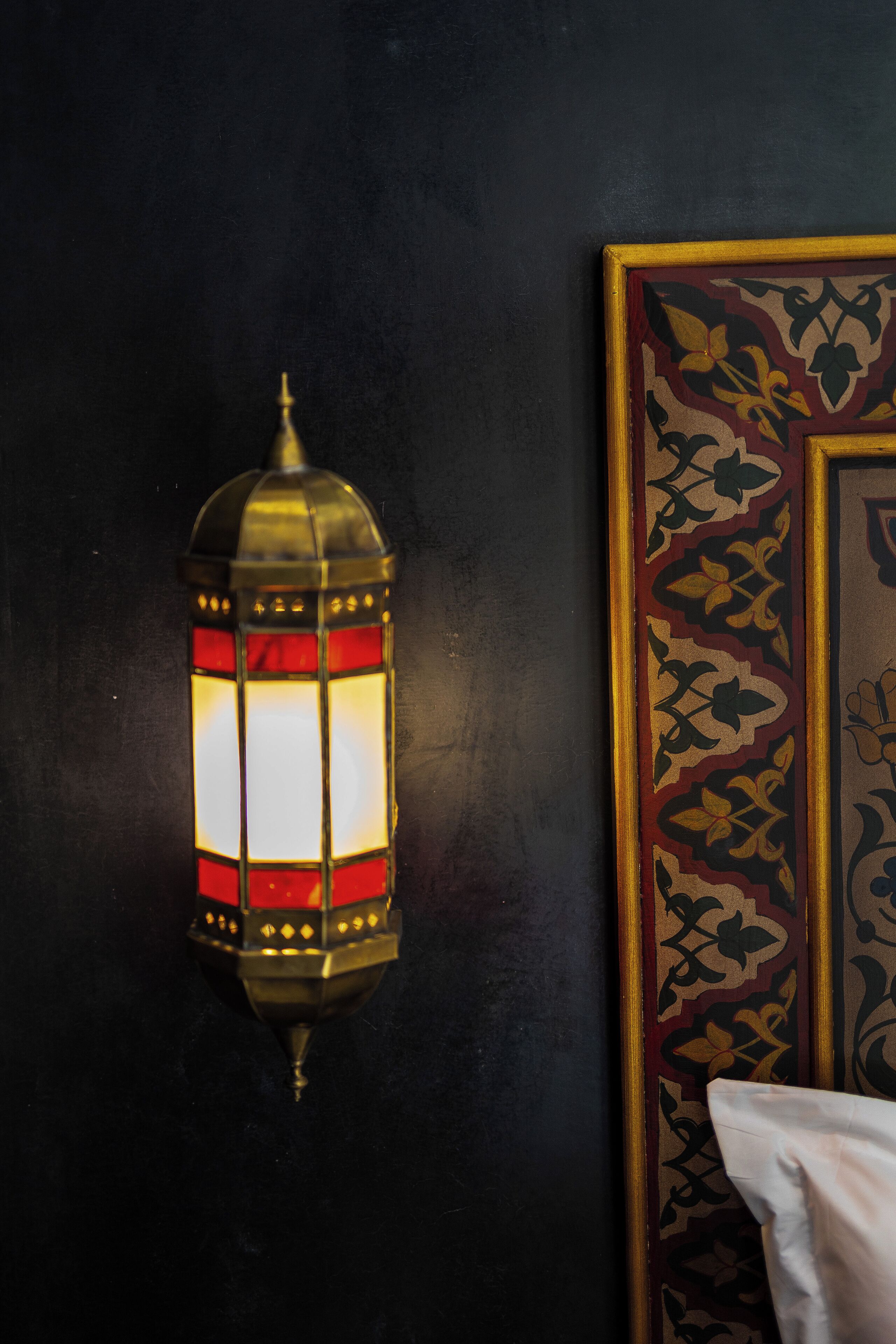 A wall-mounted lantern with a red and gold design, a framed artwork with a floral pattern, and a white pillow on a dark surface.