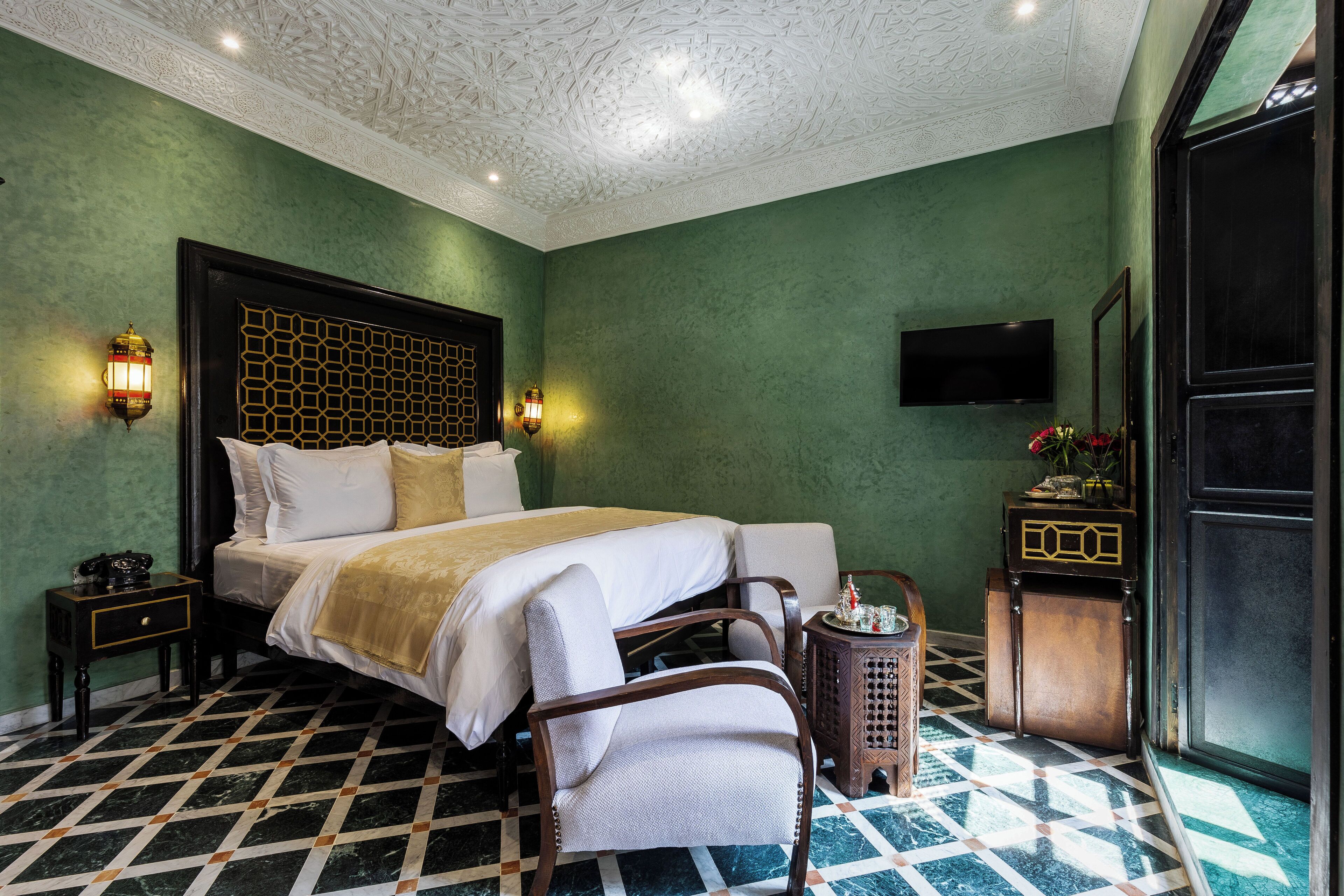 Una habitación de hotel con una cama grande, dos mesitas de noche, una lámpara de noche, una silla, una pequeña mesa con un florero y un televisor montado en la pared.