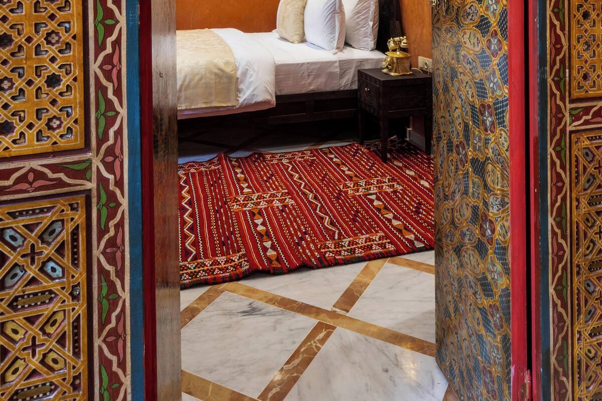 A room with a bed, a nightstand, a red patterned rug, and ornate wall decorations.