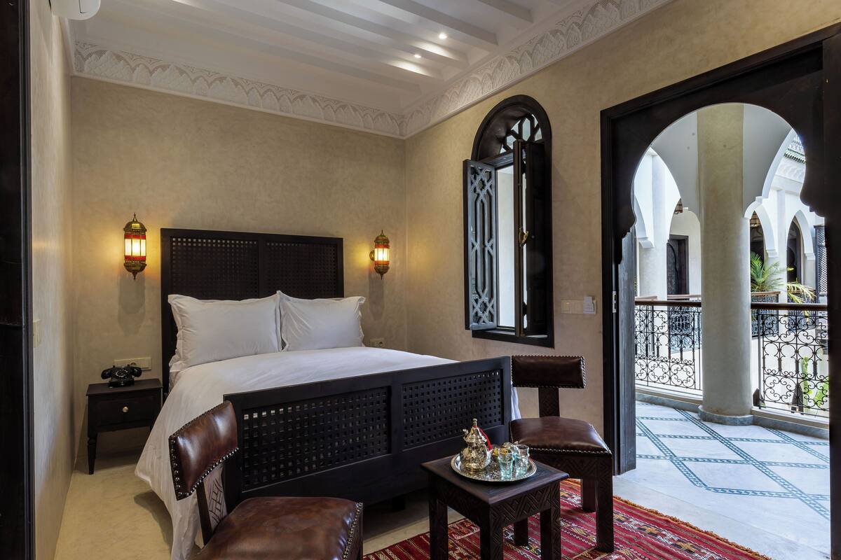A bedroom with a large bed, two bedside lamps, a nightstand, a small table with a tray, and a view of an outdoor balcony with a railing.