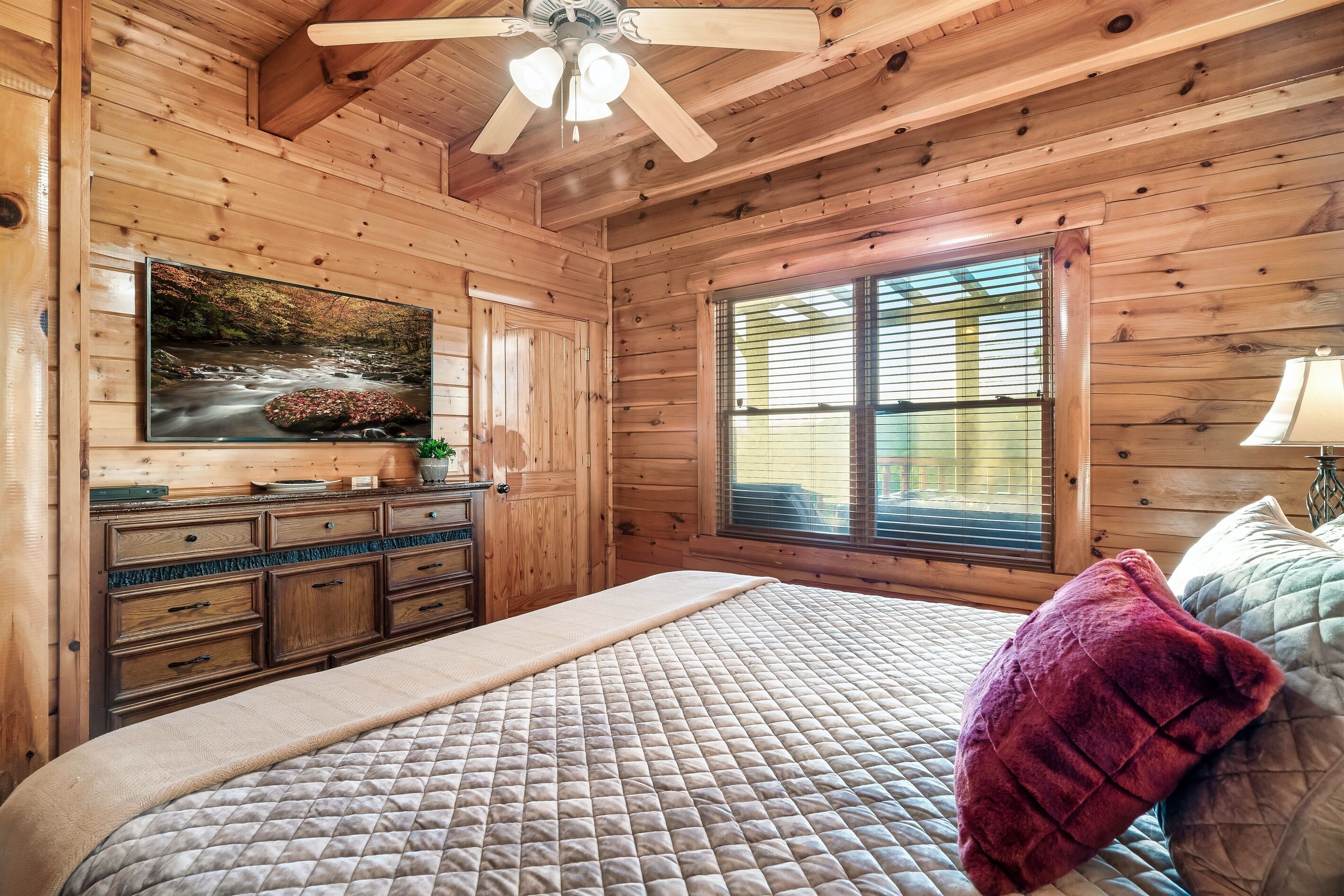 A wooden cabin bedroom with a bed, a dresser, a television, and a window.