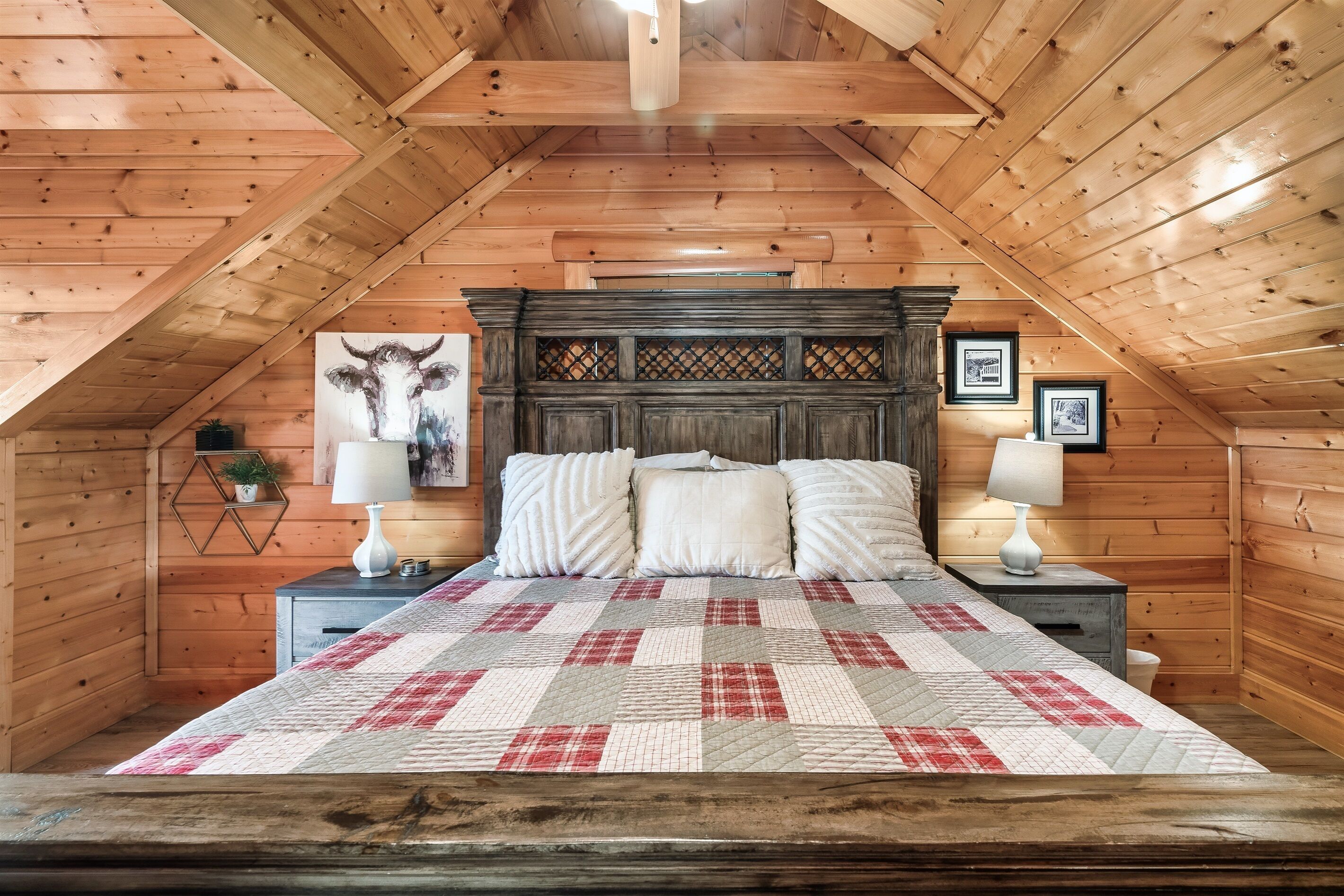 A cozy attic bedroom with a wooden sloped ceiling, a large bed with a red and white quilt, two bedside tables with lamps, and framed pictures on the walls.