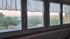 Ein Zimmer mit drei Fenstern, jedes mit weißem Vorhang und Blick auf eine Landschaft mit Bäumen und Hügeln.