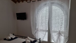 Ein Hotelzimmer mit Bett, Fernseher an der Wand und einem großen Fenster mit durchsichtigen Vorhängen.
