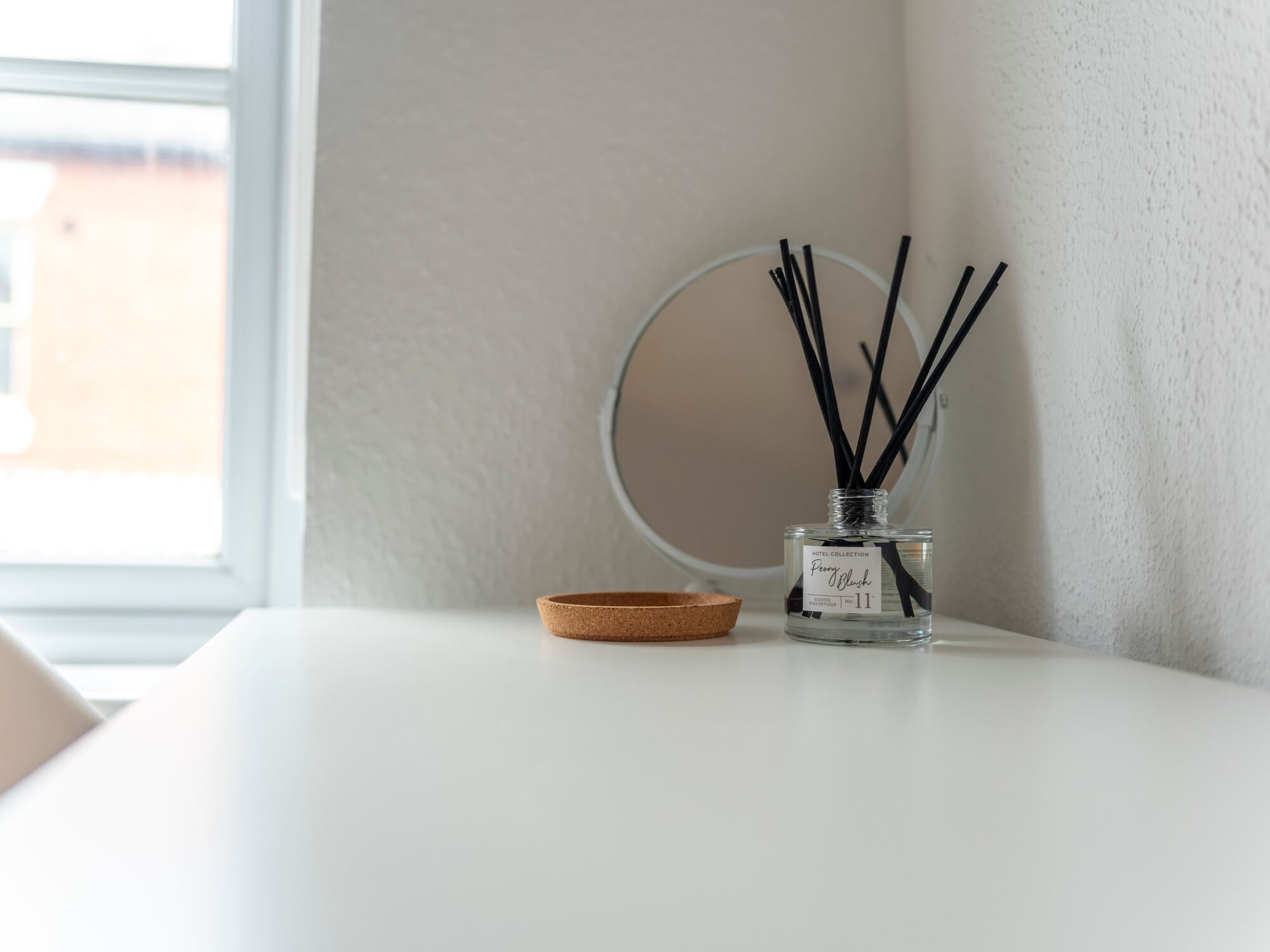A clear glass bottle with black reeds and a cork lid on a white table next to a round mirror.