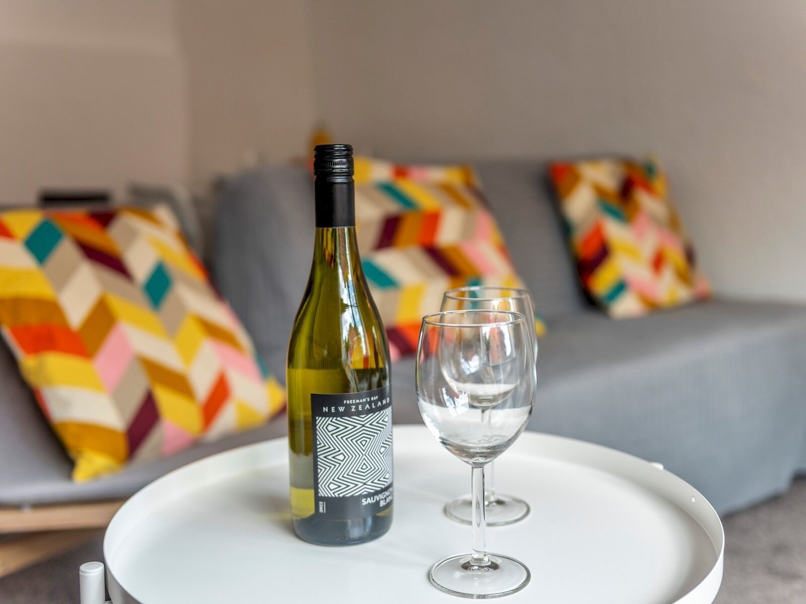 A bottle of wine and an empty glass on a tray, placed on a bed with colorful pillows.