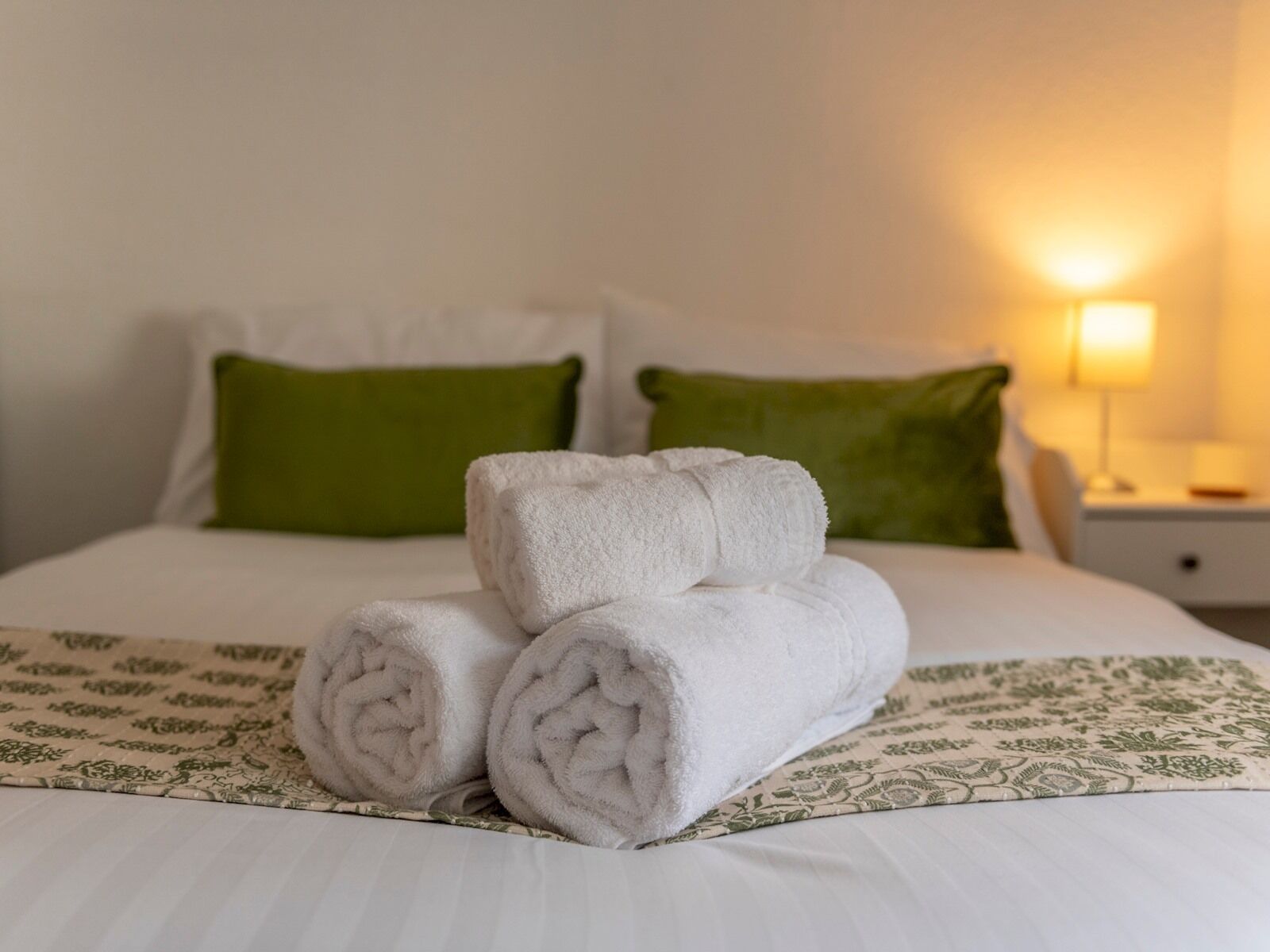 Un letto rifatto con cura, asciugamani bianchi arrotolati, cuscini verdi e una coperta a motivi.