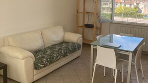 Une pièce comprenant un canapé, une table en verre avec des chaises, un téléviseur posé sur un meuble en bois et une fenêtre donnant sur la ville.