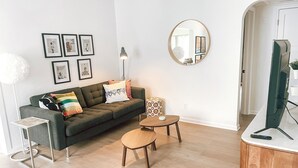 Ein modernes Wohnzimmer mit einem grauen Sofa, einem runden Spiegel, einem kleinen Holztisch und einem Flachbildfernseher.
