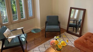 Een woonkamer met een blauwe fauteuil, een houten salontafel met bloemen, een spiegel en een kamerbreed tapijt met een patroon.